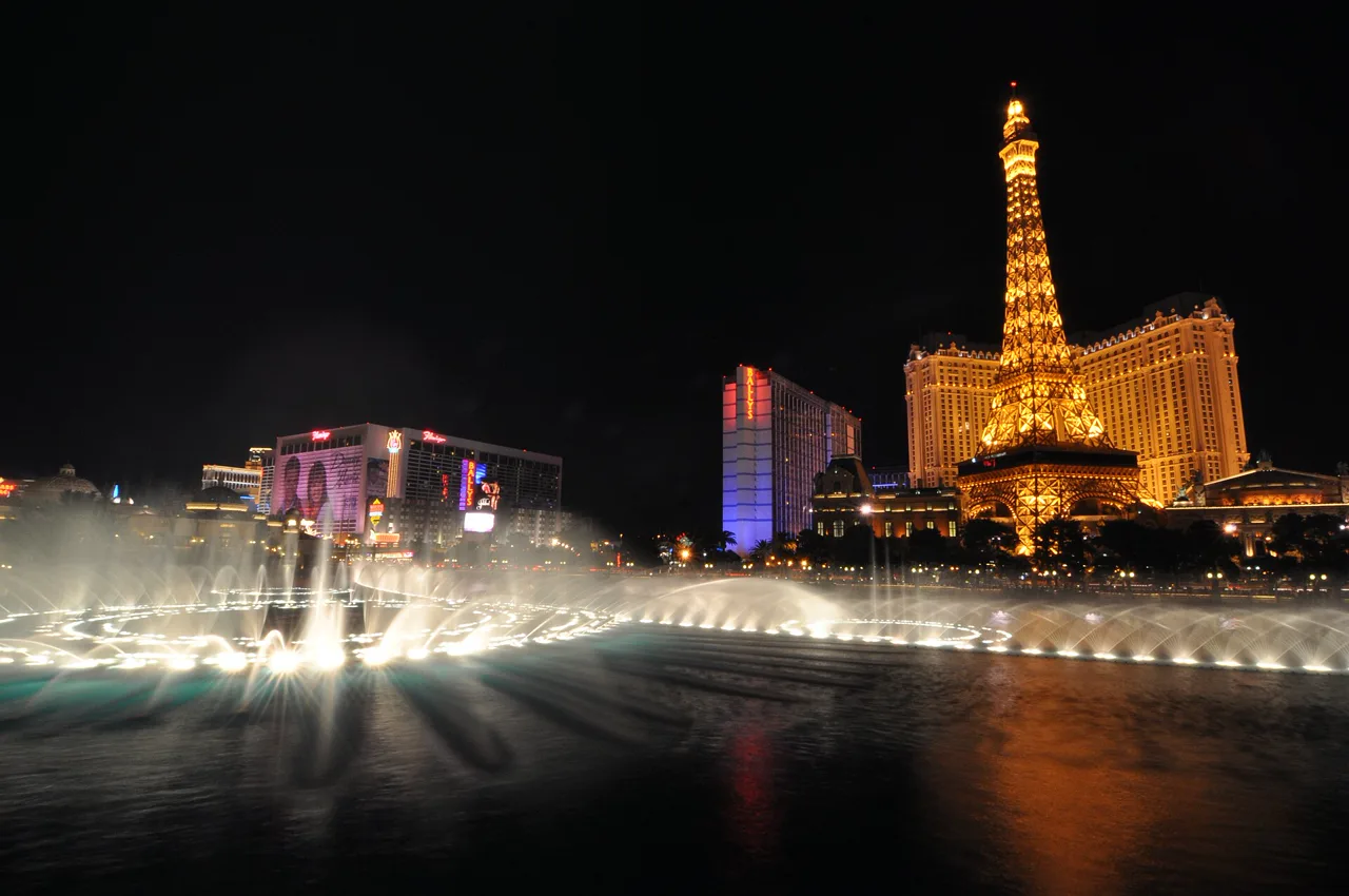 Illuminated Las Vegas fountains erupting from a lake at night with a lit Eiffel Tower replica across the boulevard