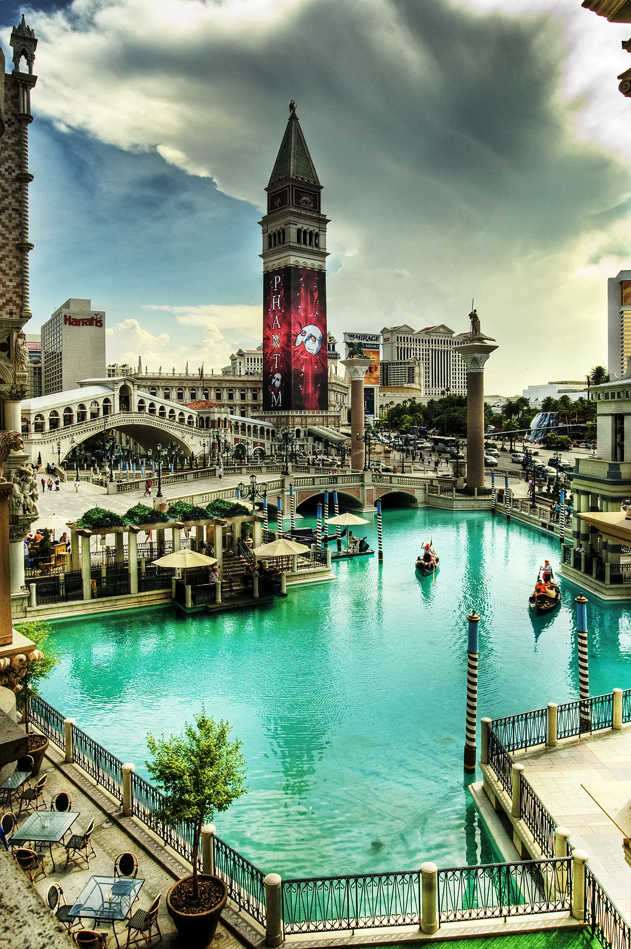 Outdoor resort canal with a tall Venetian bell tower, arched stone bridge, gondolas and turquoise water